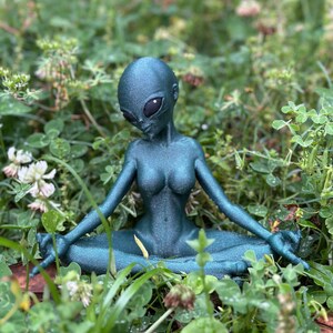 May include: A teal green alien figurine sitting in a meditative pose with its legs crossed. The figurine is sitting in a bed of green grass.