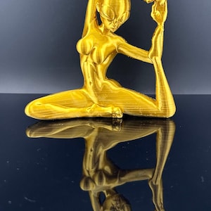 May include: A gold-colored 3D printed figurine of a person in a yoga pose. The figurine is sitting with one leg extended and the other leg bent. The person's arm is raised above their head and their hand is touching their foot.