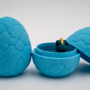 May include: A light blue, textured egg-shaped container, with a matching lid, and a small, dark green creature with a yellow crest inside. The egg is open, revealing the creature. The egg is closed in the image.