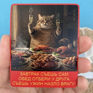 May include: A red and gold sticker with a cartoon cat holding two glasses of champagne. The cat is sitting at a table with a variety of food. The text on the sticker reads: "Breakfast you eat alone, lunch you share with a friend, dinner you eat to spite your enemy!"