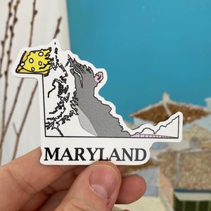 May include: A sticker of the state of Maryland shaped like a rat eating a slice of cheese. The sticker is white with black outlines and the words "MARYLAND" printed in black at the bottom.