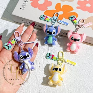 May include: A collection of Stitch character keychains in blue, pink, purple, and yellow. Each keychain features a small, plush Stitch figure with a matching colored clasp and a charm that says "Stitch".