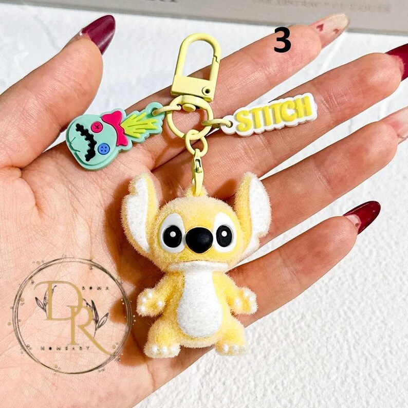 May include: A yellow Stitch keychain with a fuzzy texture, black eyes, and white accents. The keychain includes a charm with the word "STITCH" and a green and pink alien charm. The keychain is attached to a yellow metal clasp.