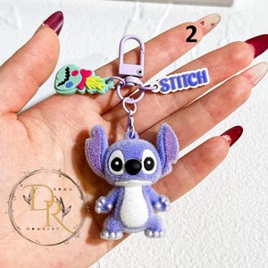 May include: A purple Stitch keychain with a fuzzy texture, featuring large black eyes and a white belly. The keychain includes a small charm and a tag that says "STITCH". The keychain is attached to a purple clasp.