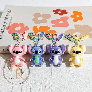 May include: Four character keychains in pink, blue, purple, and yellow. Each keychain features a cartoon character with large ears and three black eyes. The keychains have a metal clasp and a small charm with the character's name.