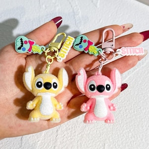 May include: Two keychains featuring cartoon character Stitch. One keychain is yellow, the other is pink. Each keychain has a matching charm with the word "Stitch" and a cartoon design. The keychains are attached to a metal clasp.