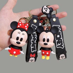Puede incluir: Llaveros con los personajes de Mickey y Minnie Mouse. Los llaveros tienen correas negras con los nombres "Minnie" y "Mickey" en blanco. El llavero de Minnie Mouse tiene un lazo rojo con lunares blancos. El llavero de Mickey Mouse tiene pantalones cortos rojos.