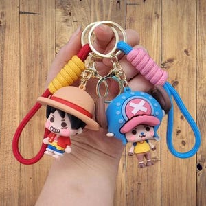 May include: Two keyrings featuring cartoon characters. One keyring has a character wearing a straw hat, red shirt, and blue shorts. The other keyring has a character with a blue hat and a pink hat. Both keyrings have gold rings and colourful cords.