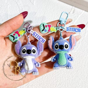 May include: Two Stitch character keychains. One is purple with a white chest and the other is blue with a green scarf. Each keychain has a matching charm and the word "STITCH" in blue letters.