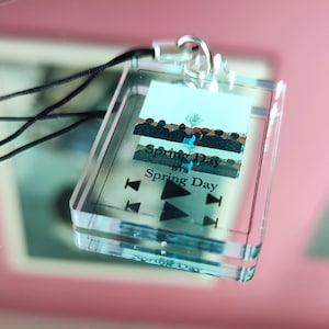 May include: A clear plastic keychain with a photo of a group of people and the text "Spring Day" and "BTS Spring Day". The keychain has a black cord and a silver ring.