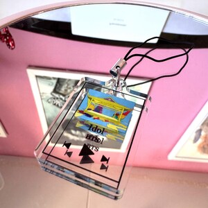May include: A clear acrylic keychain with a colorful image of a building and the text "Idol BTS" printed on it. The keychain has a black cord.