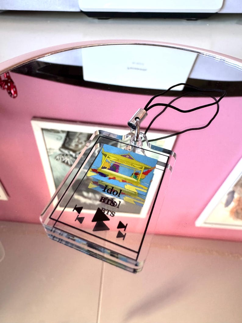 May include: A clear acrylic keychain with a colorful image of a building and the text "Idol BTS" printed on it. The keychain has a black cord.