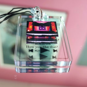 May include: A clear acrylic keychain with a black string. The keychain features a small image of a stage with red curtains and the text "How you like that?" with a play button and rewind buttons.