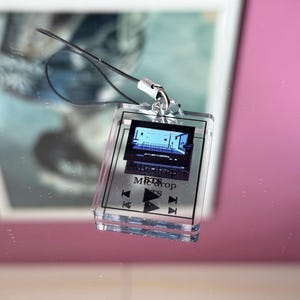 May include: A clear plastic keychain with a small screen displaying a music video. The screen is labeled "Mic Drop" and "BTS". The keychain has buttons for play, pause, and rewind.