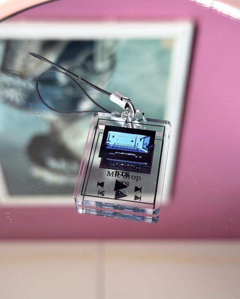May include: A clear plastic keychain with a small screen displaying a music video. The screen is labeled "Mic Drop" and "BTS". The keychain has buttons for play, pause, and rewind.