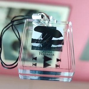 May include: A clear acrylic keychain with a black and white image of a person wearing a hat. The keychain has a black lanyard and the text "Halazia" printed on it.