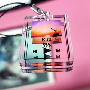 May include: A clear acrylic keychain with a colorful sunset image and the text "Run BTS Run". The keychain has a black lanyard and a silver metal ring.