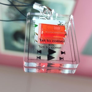 May include: A clear acrylic keychain with a red piano and the text "Yet to come" and "BTS" printed on it. The keychain has a black lanyard and a silver clasp.