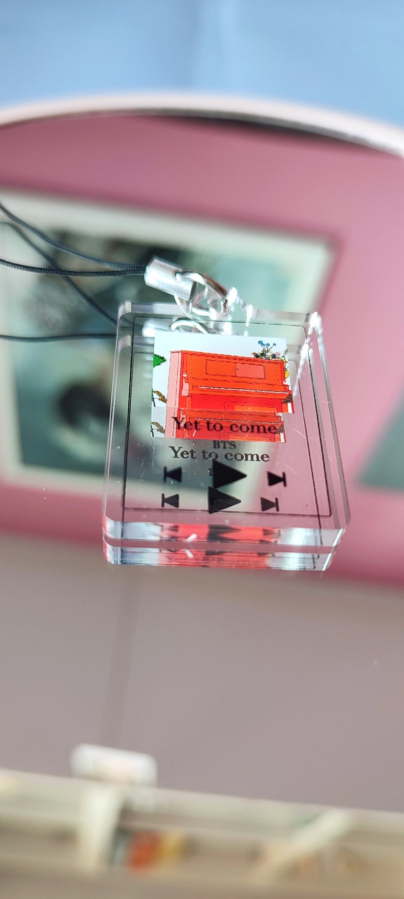 May include: A clear acrylic keychain with a red piano and the text "Yet to come" and "BTS" printed on it. The keychain has a black lanyard and a silver clasp.