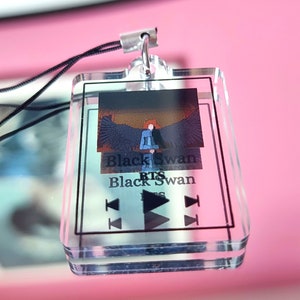 May include: A clear acrylic keychain with a black and white image of a person with wings. The text "Black Swan BTS Black Swan" is printed on the keychain.