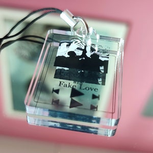 May include: A clear acrylic keychain with a black and white photo of a group of people. The keychain has a black lanyard and the text "Fake Love" printed on it.