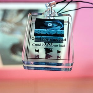 Könnte beinhalten: Ein transparenter Acryl-Schlüsselanhänger mit einem blauen und weißen Bild eines Auges und dem Text "Good boy gone bad" und "TXT" auf weißem Hintergrund. Der Schlüsselanhänger hat einen silbernen Ring und eine schwarze Schnur.