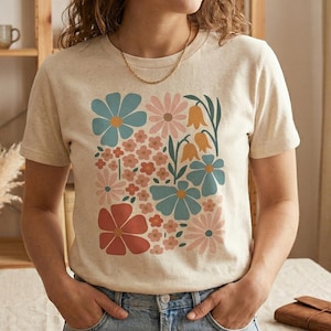 Può includere: Una maglietta beige con un design floreale colorato con fiori blu, rosa, arancioni e rossi. La maglietta è realizzata in un materiale morbido ed è indossata da una persona.