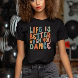 Könnte beinhalten: Ein schwarzes T-Shirt mit dem Text "Life is better when you dance" in bunten Buchstaben. Der Text ist in einer Schriftart im Retro-Stil.