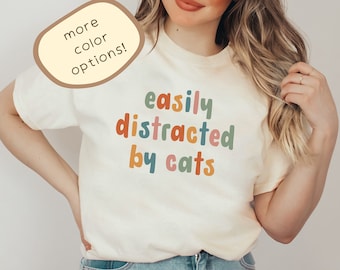 May include: A white t-shirt with the text "easily distracted by cats" in colorful letters. The text is in a playful, hand-drawn style. The model is wearing the shirt and smiling.