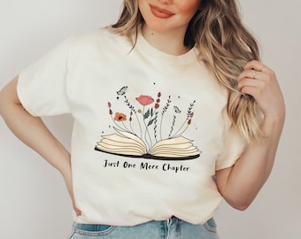 Peut inclure: T-shirt crème avec un motif de livre ouvert, des fleurs et des papillons. Le texte "Just One More Chapter" est imprimé en dessous du livre. Le t-shirt est porté avec un jean bleu.