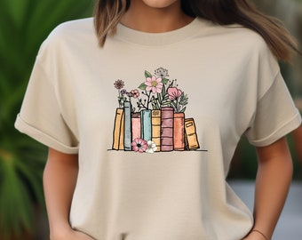 Peut inclure: T-shirt beige à col rond avec un motif de livres et de fleurs. Les livres sont de différentes couleurs, dont le jaune, le bleu, le rose et l'orange. Les fleurs sont roses et blanches.