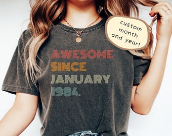 Puede incluir: Una camiseta gris oscuro con el texto "AWESOME SINCE JANUARY 1984" en letras retro de colores. El texto está superpuesto con un efecto de rayas. La camiseta la lleva puesta una persona con el pelo largo y castaño. El texto "custom month and year!" está en un bocadillo de diálogo.