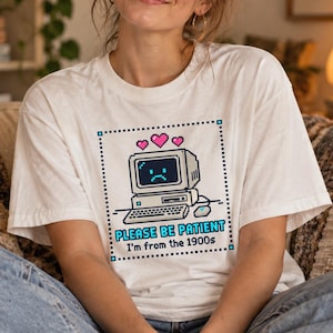 Może przedstawiać: Koszulka unisex z krótkim rękawem z grafiką pixel art przedstawiającą komputer w stylu vintage ze smutną buźką na ekranie, trzema różowymi sercami powyżej oraz napisem "PLEASE BE PATIENT I'm from the 1900s" w kropkowanej kwadratowej ramce.
