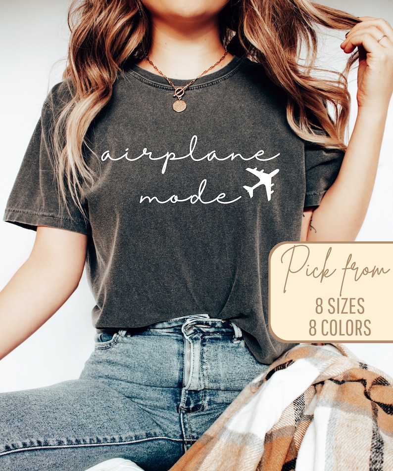 Puede incluir: Una camiseta gris oscuro con el texto "airplane mode" y un peque&ntilde;o gr&aacute;fico de avi&oacute;n en blanco. El texto "Pick from 8 sizes 8 colors" est&aacute; en un cuadro amarillo.