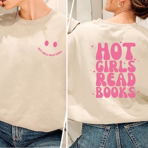 Könnte beinhalten: Beiges Sweatshirt mit dem Text "HOT GIRLS READ BOOKS" in Pink auf dem Rücken. Auf der Vorderseite befindet sich ein rosa Smiley mit dem Text "HOT GIRLS READ BOOKS" in Pink. Das Sweatshirt hat einen lässigen Stil.