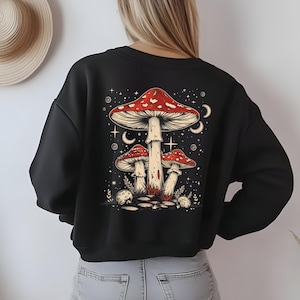 Könnte beinhalten: Schwarzes Sweatshirt mit einer farbenfrohen Illustration von drei rot-weißen Pilzen vor einem Sternenhimmel mit Monden und Sternen. Das Sweatshirt hat einen Rundhalsausschnitt.