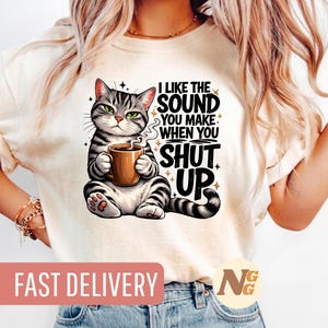 Puede incluir: Camiseta color crema con un gato de dibujos animados sosteniendo una taza de café. El gato es de rayas grises y negras con ojos verdes. El texto en la camiseta dice "I LIKE THE SOUND YOU MAKE WHEN YOU SHUT UP."