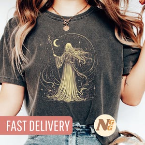 Op de afbeelding: Een donkergrijs T-shirt met een grafische print van een vrouw met lang, golvend haar die voor een halve maan en sterren staat. De tekst "SNELLE LEVERING" is gedrukt op een roze rechthoek onderaan de afbeelding.