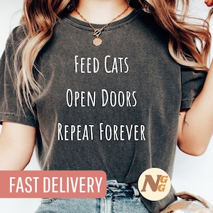 Könnte beinhalten: Ein dunkelgraues T-Shirt mit dem Text "Feed Cats Open Doors Repeat Forever" in weiß gedruckt. Das Shirt wird von einer Person mit langen braunen Haaren getragen. Der Text "FAST DELIVERY" ist in weiß auf einem rosa Rechteck am unteren Rand des Bildes gedruckt. Ein braunes und goldenes Logo befindet sich in der unteren rechten Ecke des Bildes.