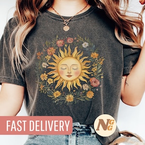 Op de afbeelding: Een donkergrijs T-shirt met een bloemenontwerp met een lachende zon met gesloten ogen. De zon wordt omringd door bloemen in tinten geel, oranje, rood en roze. De tekst "SNELLE LEVERING" is zichtbaar onderaan de afbeelding.