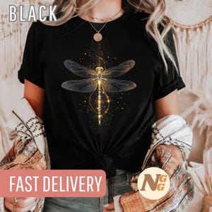 Könnte beinhalten: Schwarzes T-Shirt mit einem goldenen Libellen-Motiv und dem Text "SCHNELLER VERSAND" auf einem rosa Banner. Die Libelle ist von einem funkelnden Effekt umgeben.