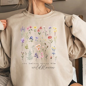 Könnte beinhalten: Ein beiger Pullover mit einem Blumenmuster und dem Text "you belong among the wildflowers".
