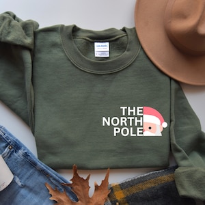 Könnte beinhalten: Ein grünes Sweatshirt mit einer weißen Grafik, auf der "The North Pole" steht, wobei das "O" in "Pole" durch ein Gesicht des Weihnachtsmanns ersetzt wird.