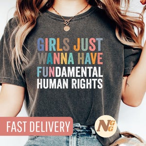 Könnte beinhalten: Ein dunkelgraues T-Shirt mit dem Text "Girls Just Wanna Have Fundamental Human Rights" in bunten Buchstaben.