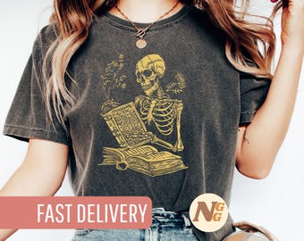 May include: A dark gray t-shirt with a graphic of a skeleton reading a book. The skeleton is drawn in a vintage style with yellow outlines. The book is open and has the word "BOOK" written on the cover.