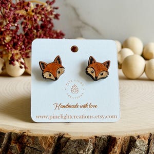 May include: A pair of stud earrings shaped like fox heads, crafted from wood with orange and brown details. The earrings are attached to a white display card featuring the text Pine Light Creations, Handmade with love, and www.pinelightcreations.etsy.com.
