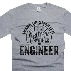 Puede incluir: Camiseta gris con un diseño gráfico negro que dice "Wake Up Smarter Sleep With An Engineer". El gráfico es un círculo con varios símbolos de ingeniería.