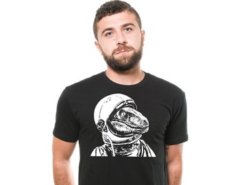 Può includere: Una maglietta nera con una grafica bianca di un velociraptor che indossa un casco da astronauta. Il raptor guarda a destra.