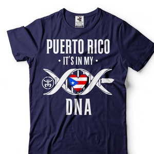 Puede incluir: Una camiseta azul marino con un diseño gráfico blanco. El gráfico presenta una cadena de ADN con un corazón en el medio. El corazón es la bandera de Puerto Rico. El texto "Puerto Rico" está en la parte superior del gráfico y el texto "It's in my DNA" está debajo del gráfico.