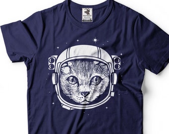 以下が含まれることがあります： 宇宙飛行士のヘルメットをかぶった猫の白いグラフィックがプリントされたネイビーブルーのTシャツ。猫の目は直接見ています。背景は濃い青色で白い星が散りばめられています。